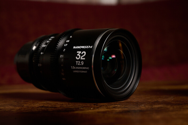 Anamorphic cinema lens with 32mm focal length and T2.9 aperture on wooden surface against red backdrop.