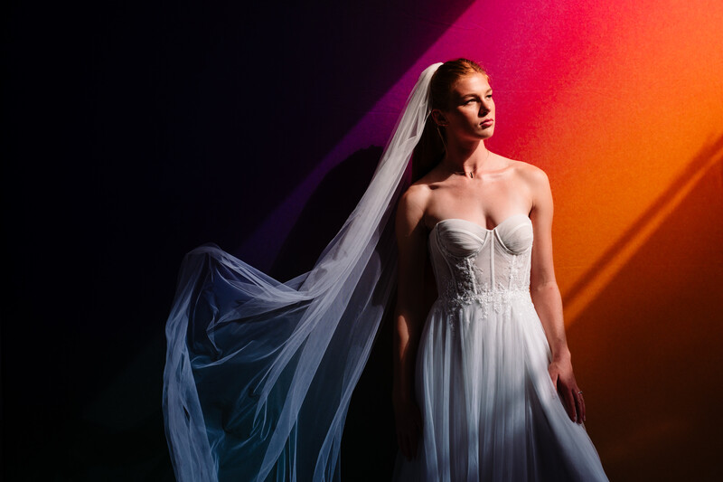 Woman in flowing white gown with veil against vibrant pink and orange gradient backdrop.