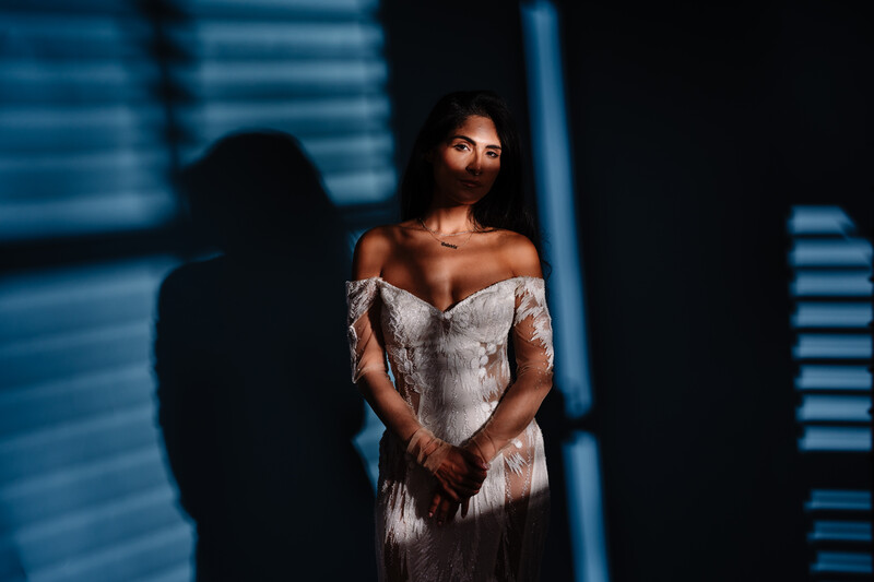 Woman in white off-shoulder dress standing against blue neon-lit background with shadow cast on wall.