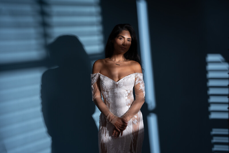 Woman in white off-shoulder dress standing against blue-toned architectural background with dramatic shadow.