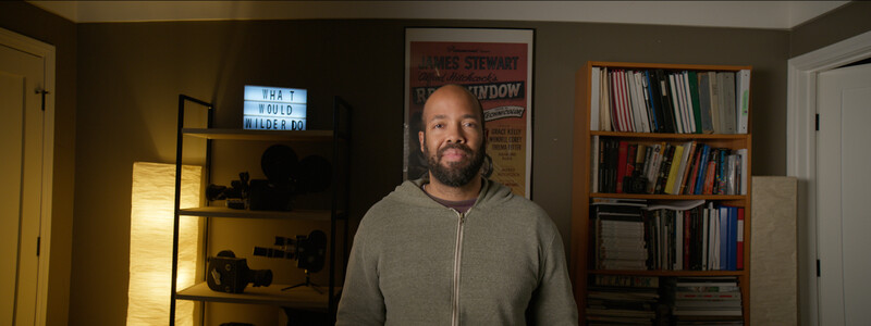 Man in gray hoodie standing in home office with bookshelves, vintage movie poster, and neon sign visible behind him.