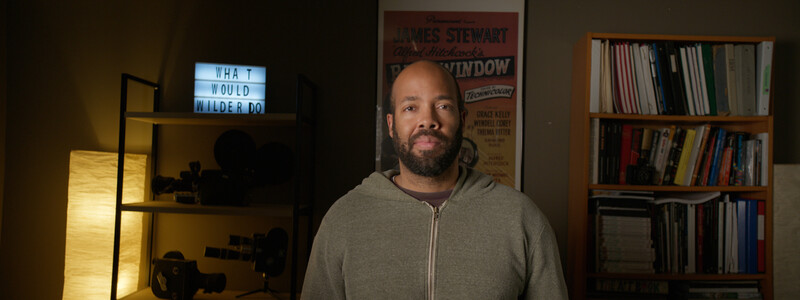 Bearded man in gray zip-up hoodie standing in a home studio with warm lighting and shelving.