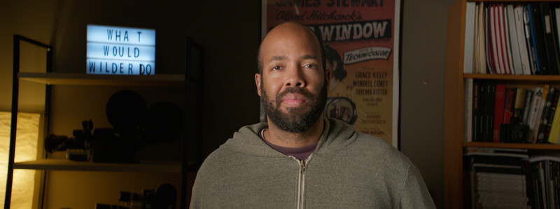 Man with beard wearing gray zip-up hoodie in a home office setting with warm lighting.