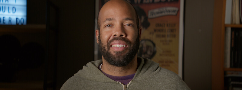 Smiling man with beard wearing a gray hoodie and purple shirt indoors.
