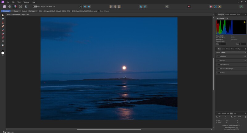 Full moon rising over calm ocean water at twilight with blue sky and dark rocky foreground.