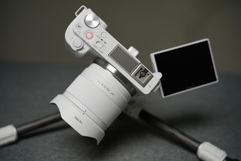 Mirrorless camera with white lens mounted on tripod, articulated LCD screen extended toward camera.