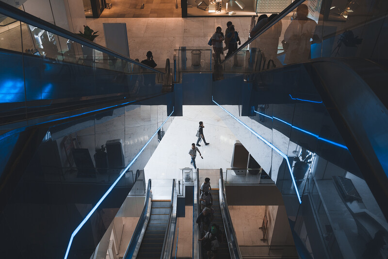 Modern multi-level interior with people on escalators and walkways, illuminated by blue accent lighting.