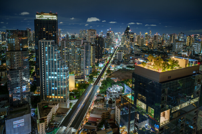 Urban cityscape at dusk with illuminated skyscrapers and a light trail from traffic on a major avenue.