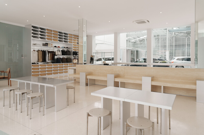 Modern minimalist retail or café interior with light wood tables, white stools, and wall-mounted shelving displaying products.