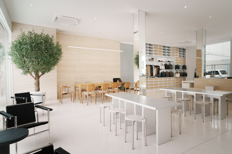 Minimalist modern office interior with white flooring, natural wood furniture, and abundant natural light from floor-to-ceiling windows.