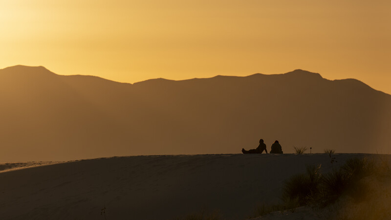 Silhouetted figures sitting on a desert dune at golden hour with mountains in the distance.