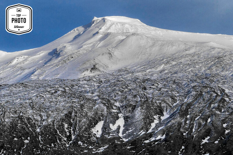 Snow-covered volcanic cone with glaciated ridges and clear blue sky.
