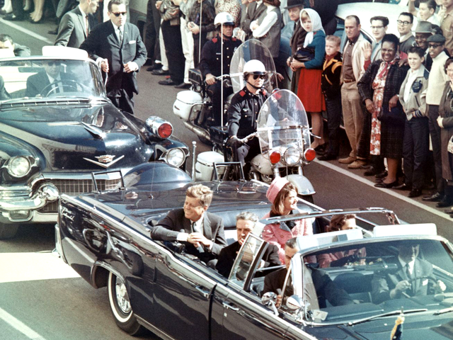 Presidential motorcade with convertible limousine traveling through crowded city street lined with spectators.