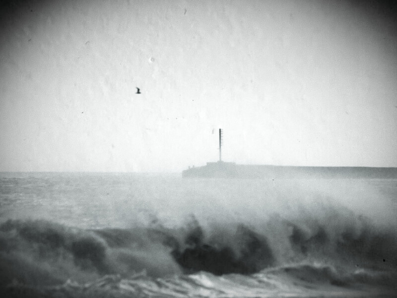Dramatic seascape with large waves crashing in foreground and a distant lighthouse on rocky headland shrouded in mist.