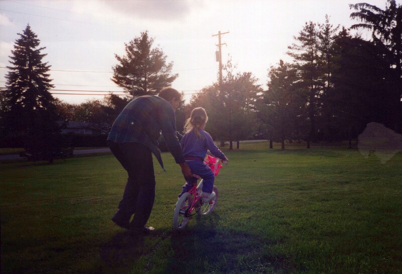 Adult bending down to help a child on a small bike in a grassy park at golden hour.