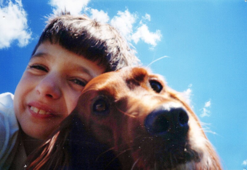 Child smiling at camera with dog's face close beside them against blue sky.