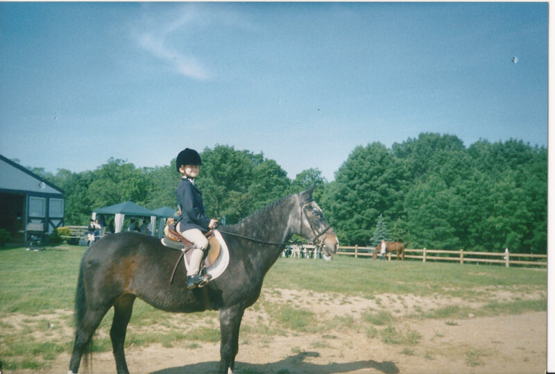 Young rider mounted on dark horse in saddle at rural equestrian facility with white fencing.