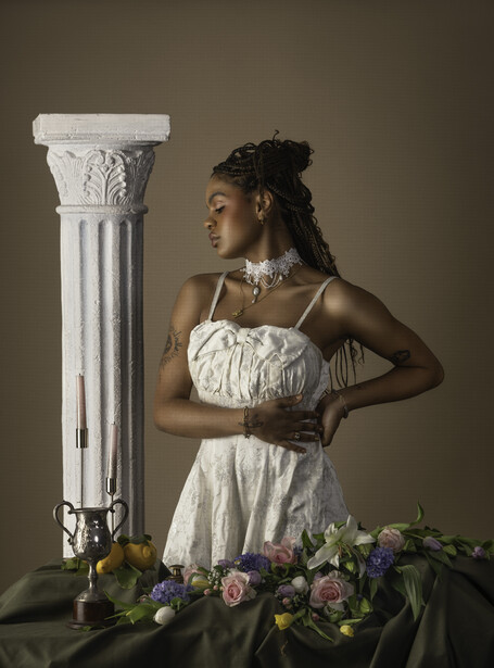 Woman in white draped dress posed beside classical white column with floral arrangement below.
