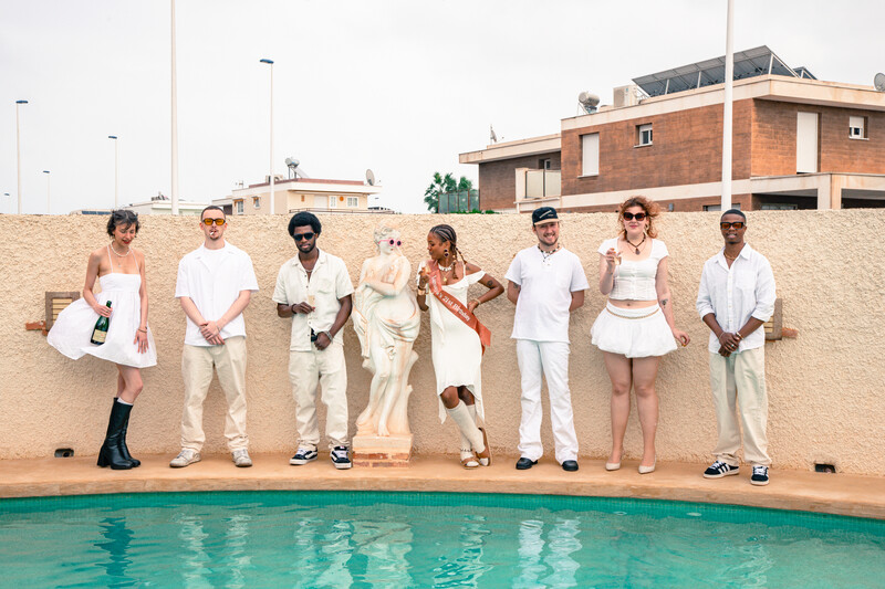 Eight people in white and neutral clothing standing in a line beside a turquoise pool against a tan concrete wall.