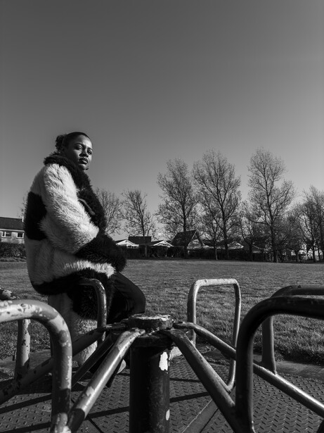 Man in fur-collared coat seated on playground equipment against bare winter trees.