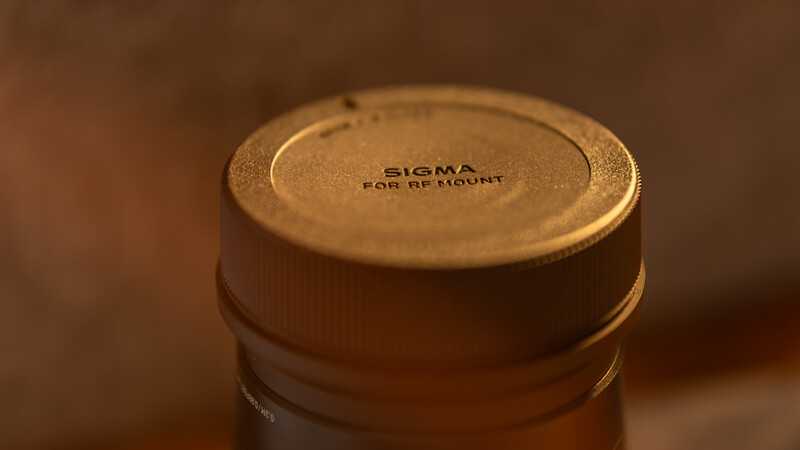 Close-up of a gold lens cap marked "SIGMA" and "FOR RF MOUNT" against a warm brown background.