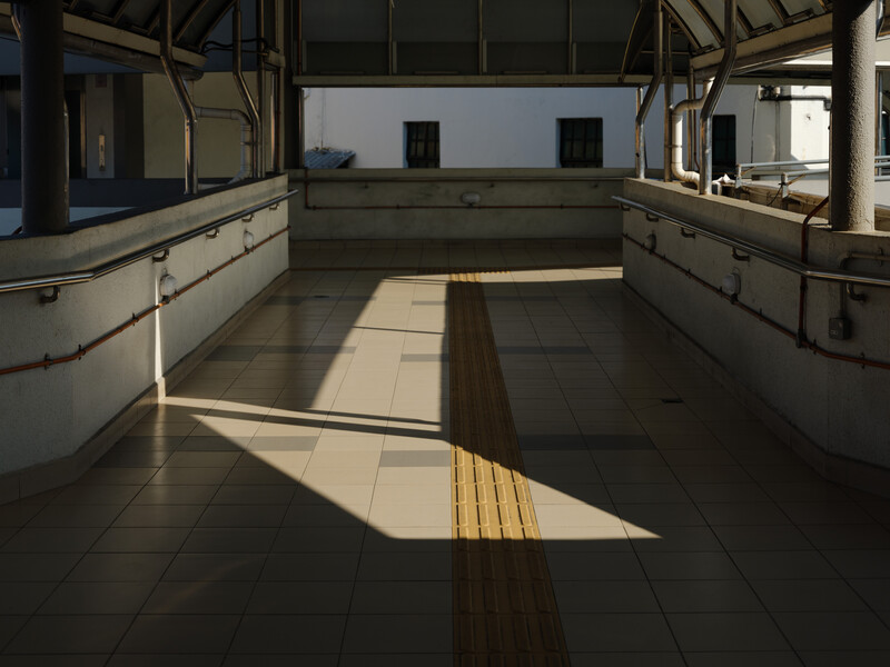 Empty train or transit platform with yellow tactile warning strip running down the center, metal handrails on both sides, and overhead structural supports casting geometric shadows.