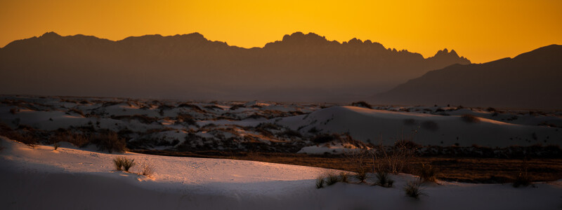 Desert landscape at sunset with golden sky and silhouetted mountains behind white sand dunes.