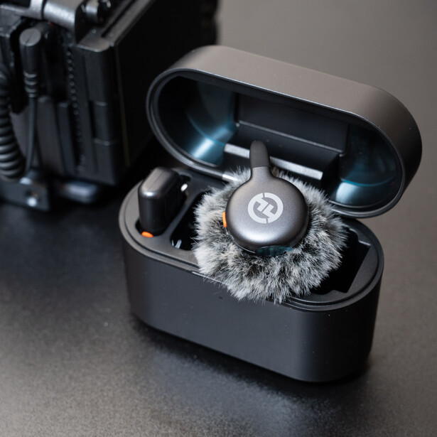 Open charging case with wireless earbuds and microphone windscreen on dark surface next to camera equipment.