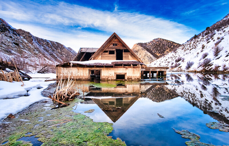 Abandoned wooden house reflected in still alpine lake surrounded by snow-covered mountains.