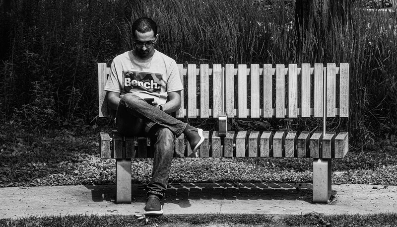 Man in glasses and bench t-shirt reading a book while seated on a concrete bench against a white picket fence.