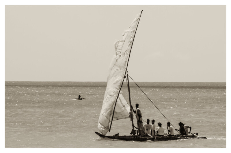 Traditional sailing vessel with white sail and crew members aboard on calm water.