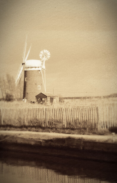 Sepia-toned landscape featuring an old windmill beside a canal with a horse grazing in the foreground.