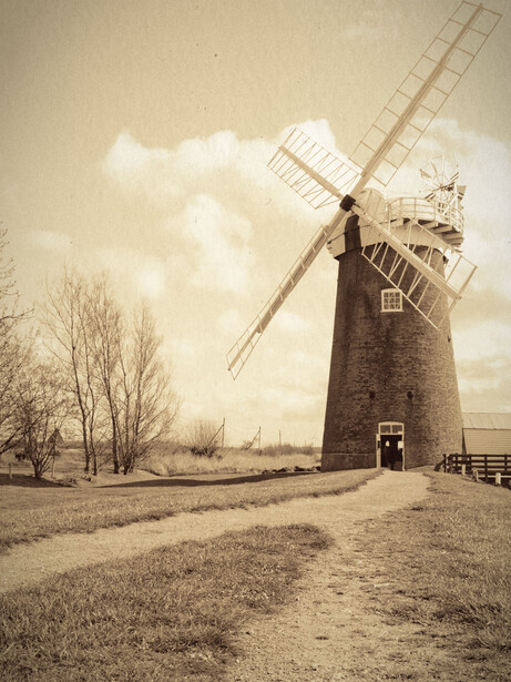 Historic brick windmill with white sails against a cloudy sky, surrounded by bare winter trees and open grassland.