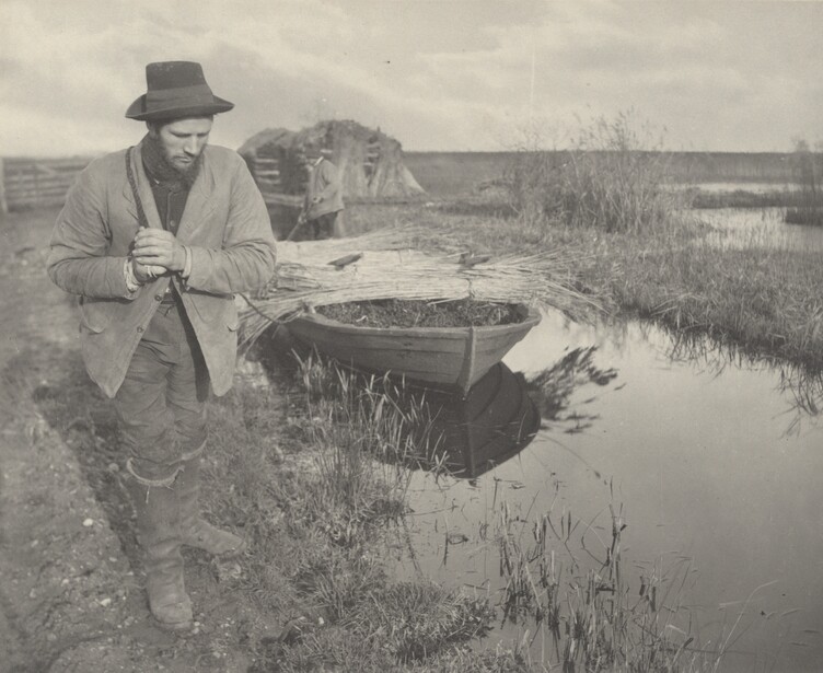 Man in hat and vest holding a large fish beside a boat in shallow wetland water.