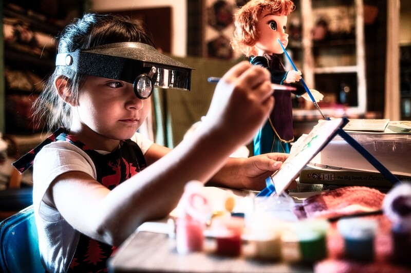 Child wearing jeweler's loupe and apron paints at table with art supplies while another child works in background.