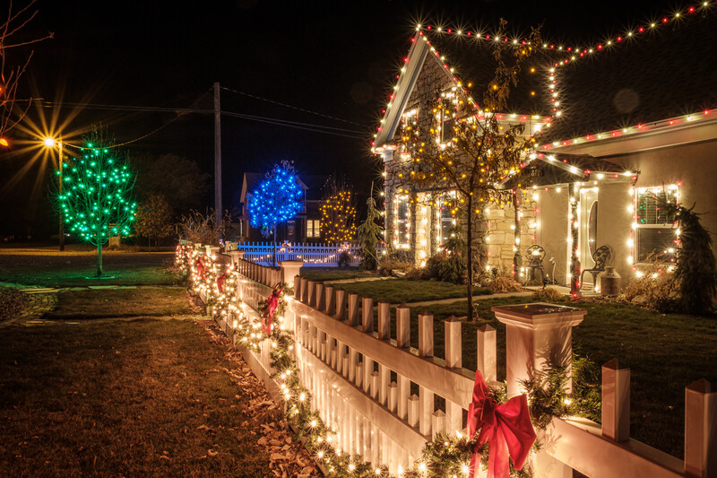 Residential home decorated with warm white and colorful Christmas lights strung across trees, fence, and roofline at night.