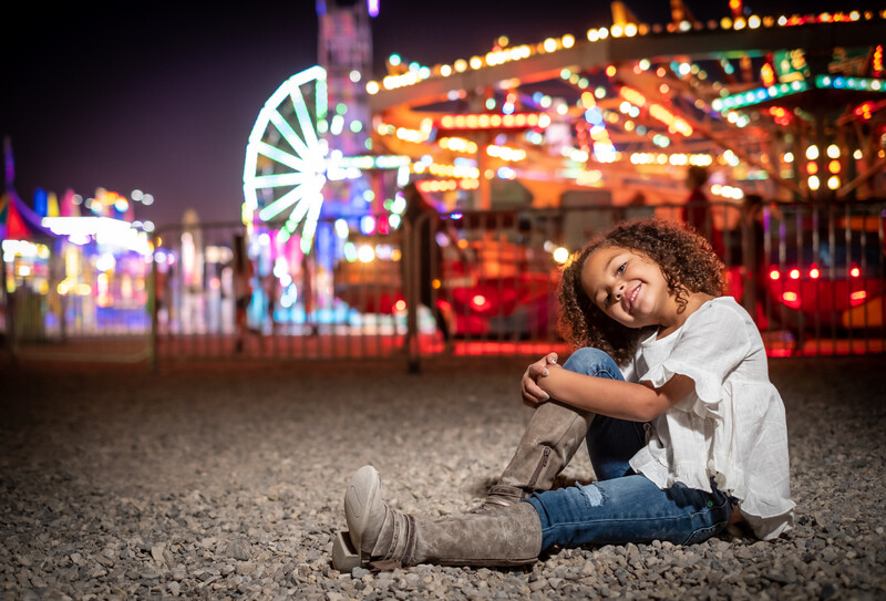 Young person sitting on sand at night with illuminated carnival rides blurred in background.