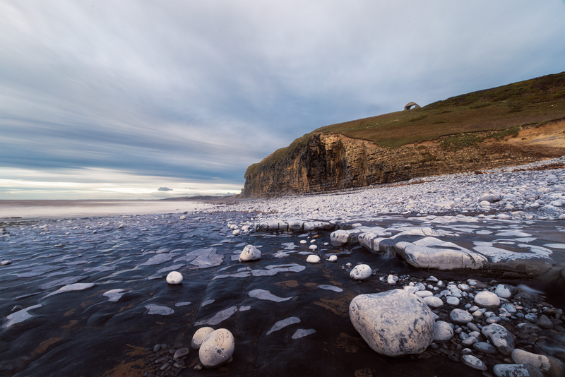 Rocky coastline with snow-covered boulders in shallow water beneath a steep cliff face at dusk.
