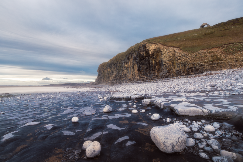 Icy rocky shoreline with snow-covered cliff face and partially frozen water at dusk.