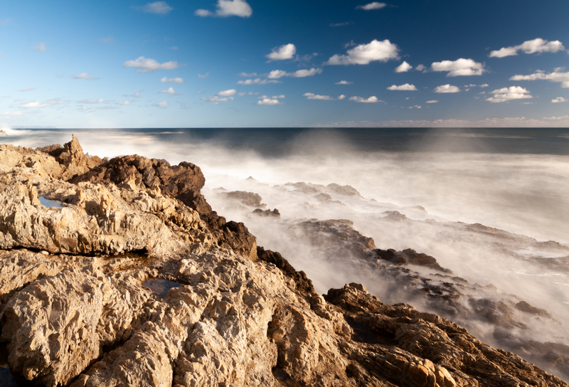 Rocky coastline with long-exposure waves creating misty white water against tan cliff formations and calm blue sky.
