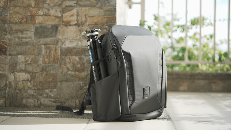 Gray and black camera backpack with tripod attachment leaning against stone wall indoors.