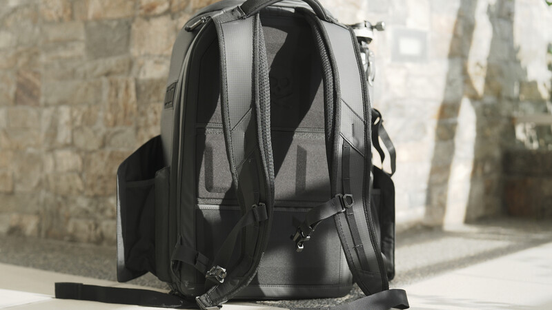 Dark gray camera backpack displayed from behind, showing padded shoulder straps and ventilated mesh panels against a stone wall.
