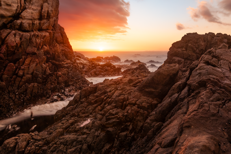 Rocky coastal landscape with dramatic sunset and misty waves flowing between large boulders.