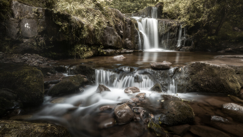 Multi-tiered woodland waterfall cascading over moss-covered rocks into a pool surrounded by boulders.