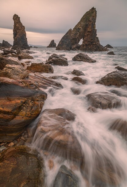 Dramatic rocky sea stacks rising from turbulent ocean waters at dusk, with smooth water motion blur in foreground.