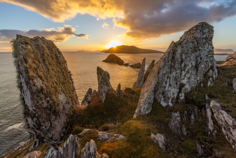 Dramatic rocky pinnacles frame a golden sunset over a calm sea with distant islands.