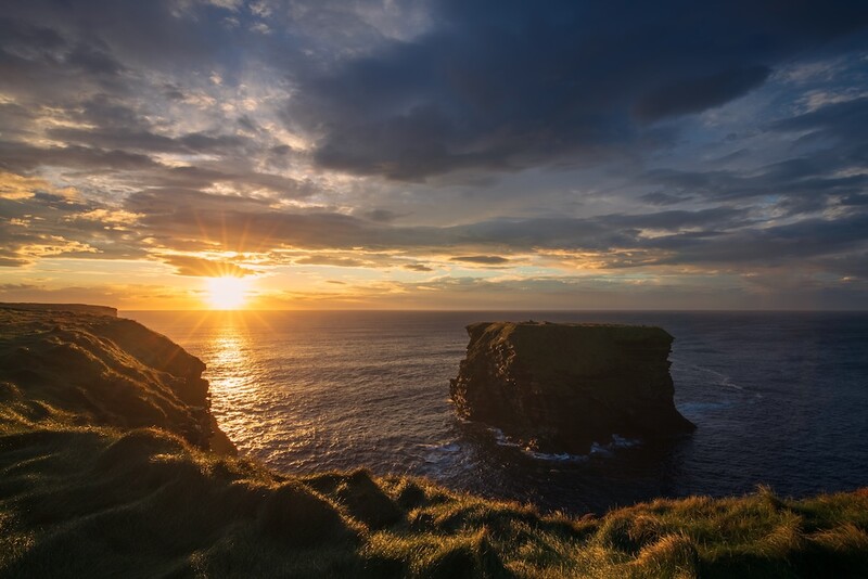 Dramatic coastal sunset with rocky sea stacks silhouetted against golden sky and ocean.