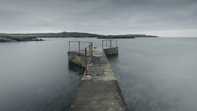 Concrete pier with metal railings extending into calm gray water toward distant rocky coastline.