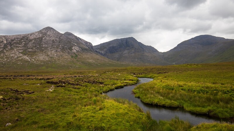 Meandering stream through moorland with three mountains under overcast sky.