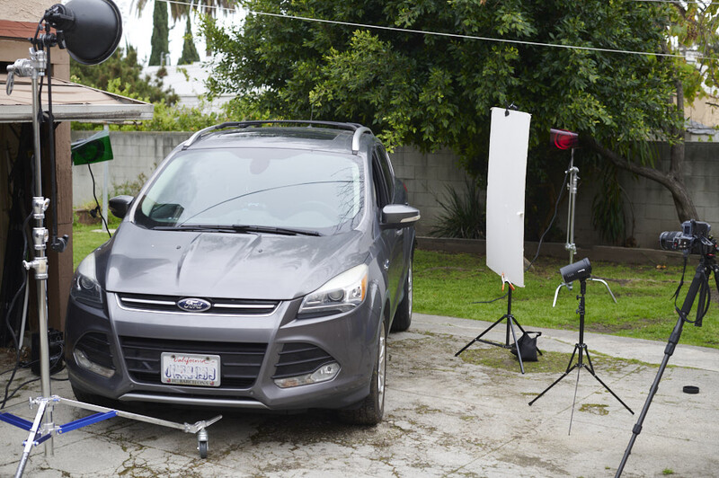 Professional product photography setup with a gray SUV lit by studio strobes and reflectors in a residential driveway.